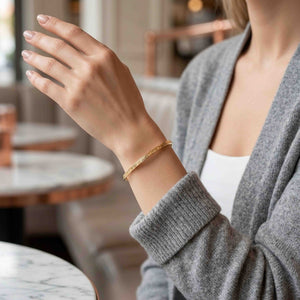 4 of 10: Close-up of a woman’s wrist wearing a slim 3.5mm linen bronze cuff bracelet with a soft hammered texture. The warm golden tone complements a casual outfit with a gray knit sweater and white top, set against a softly blurred indoor background with neutral tones.