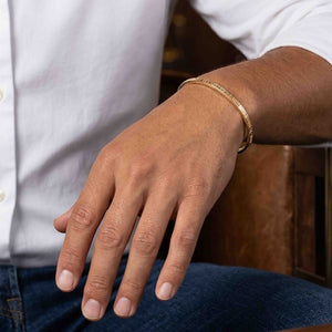 2 of 10: Close-up of a man’s wrist wearing a slim 3.5mm linen bronze cuff bracelet with a subtle hammered texture. The warm golden tone contrasts softly against the skin, styled with a white shirt and blue jeans for a casual, everyday look.