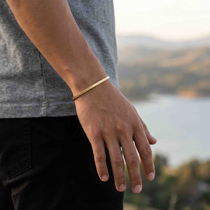 6 of 10: Close-up of a person’s wrist wearing a slim 3.5mm linen bronze cuff bracelet with a subtle hand-hammered texture. The warm golden tone stands out against a casual outfit with a gray shirt and black pants, set outdoors with a softly blurred landscape and water in the background.