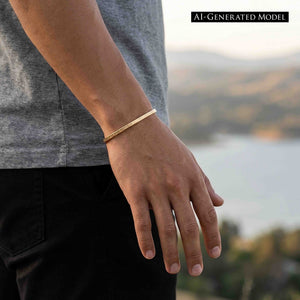 6 of 10: Close-up of a person’s wrist wearing a slim 3.5mm linen bronze cuff bracelet with a subtle hand-hammered texture. The warm golden tone stands out against a casual outfit with a gray shirt and black pants, set outdoors with a softly blurred landscape and water in the background.  AI Generated Model.