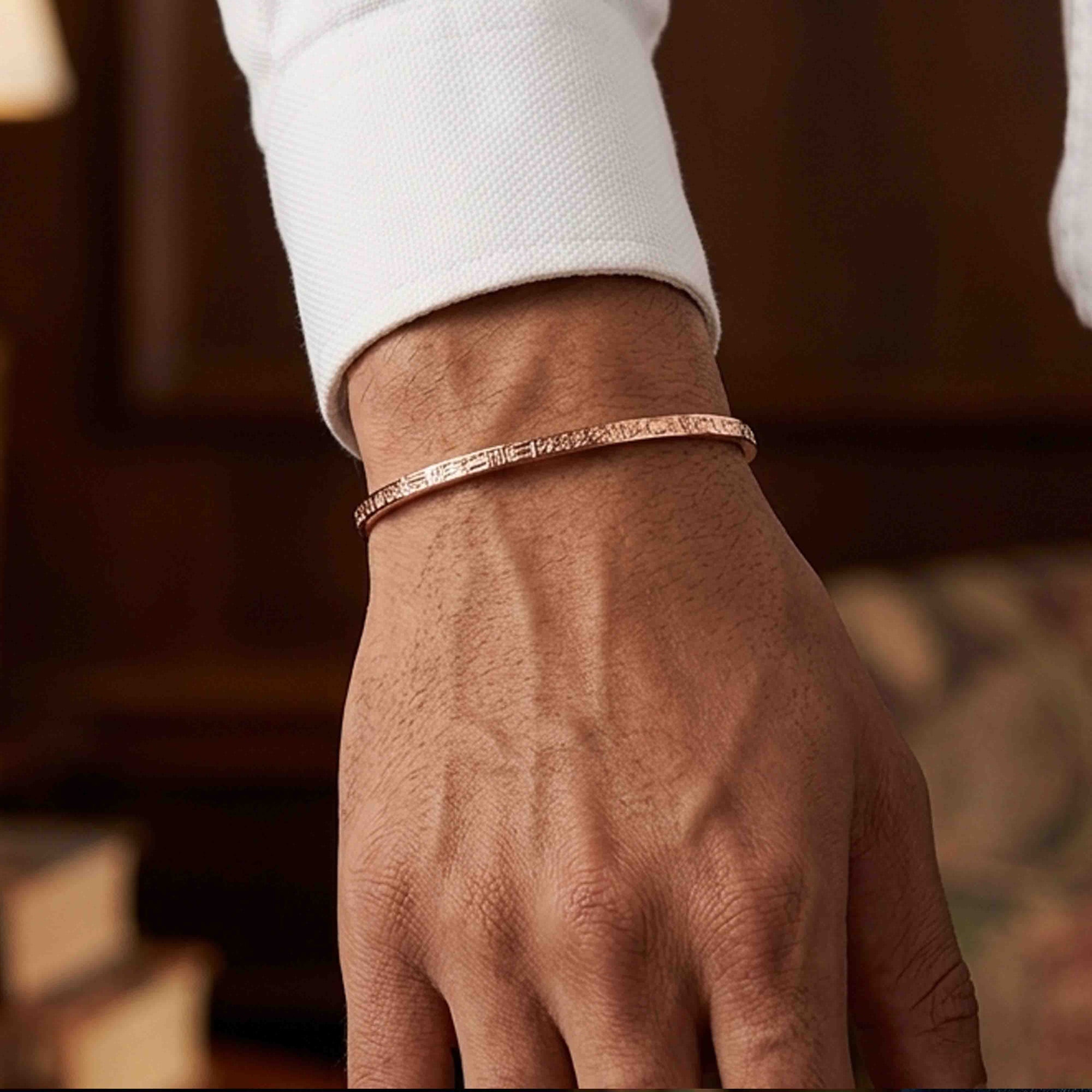 Close-up of a hand wearing a slim solid copper cuff bracelet with a subtle linen-style textured surface. The narrow bracelet sits neatly at the wrist below a white dress shirt cuff, giving it a clean, understated look. Warm indoor lighting highlights the copper’s soft rose tone, while the blurred wood-paneled background adds a refined, classic setting suited to both casual and dress wear.