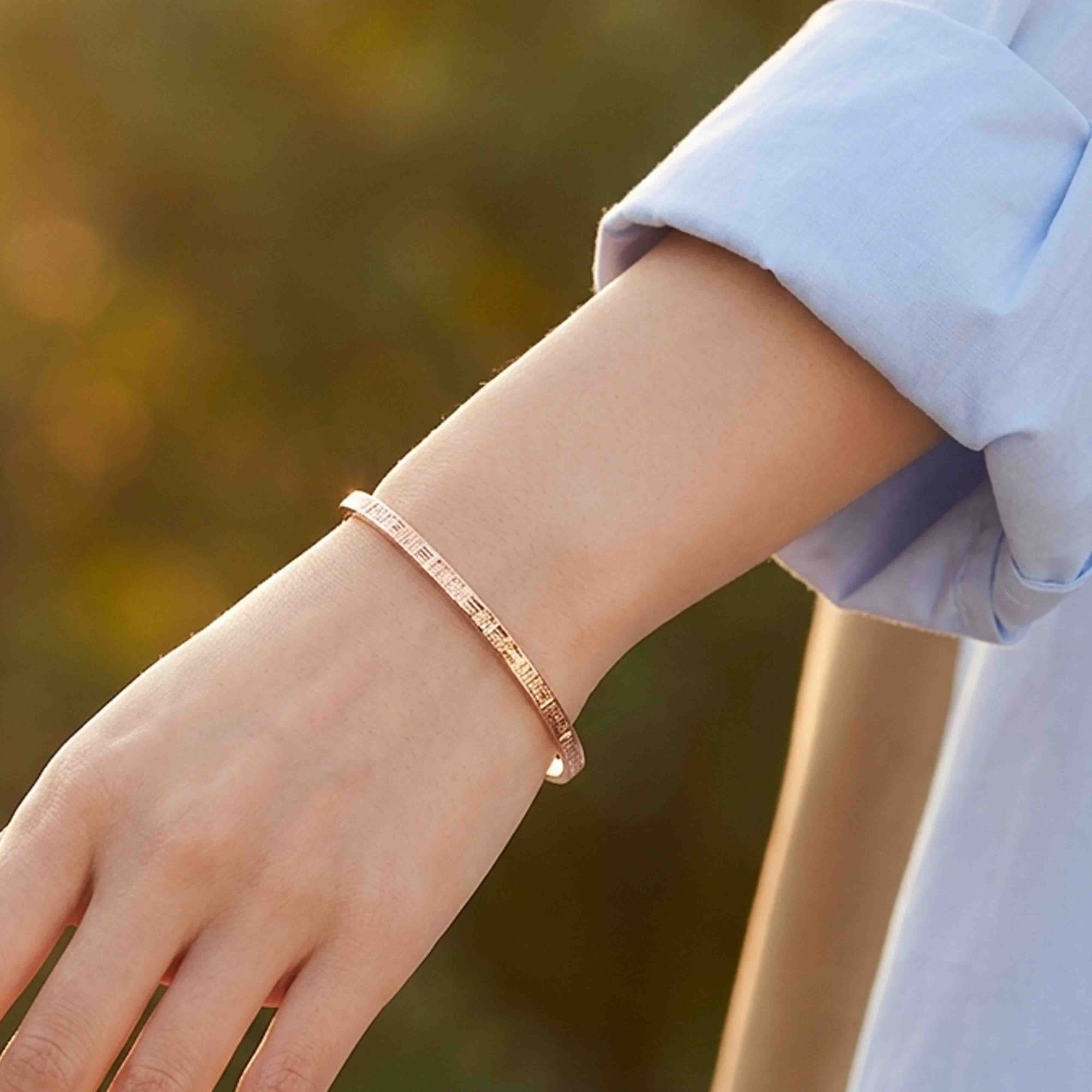 Woman wearing a slim 3.5mm Linen Copper Cuff Bracelet on her wrist with a light blue rolled-sleeve shirt. The narrow open bracelet features a warm polished copper finish and fine linear texture. The close-up outdoor photo highlights the delicate handmade design against a softly blurred green and golden background.