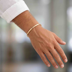 6 of 9: Man wearing a narrow matte bronze cuff bracelet with a white shirt sleeve. The handcrafted solid bronze cuff has a clean minimalist design with a brushed matte finish and warm golden bronze color, creating a modern unisex jewelry look for everyday style.