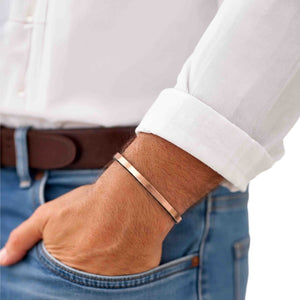 8 of 10: Matte copper cuff bracelet worn on a man’s wrist with a rolled white shirt sleeve, showing its narrow profile, warm copper tone, and casual polished style.