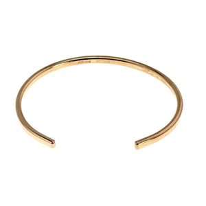 9 of 10: Front product view of a narrow polished bronze cuff bracelet on a white background. The open ends face forward at the bottom, while a faint John S. Brana bronze mark appears along the inside top edge of the warm golden band.