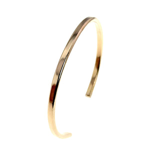 5 of 10: Angled side product view of a narrow polished bronze cuff bracelet standing upright on a white background. The curved band and open construction are clearly visible, along with its smooth edges, rounded end, slim profile, and bright golden finish.