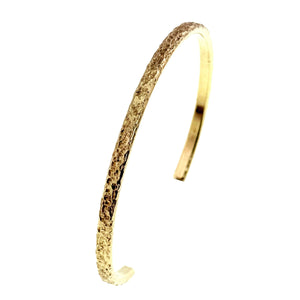 7 of 9: Close angled side view of a slim Nu Gold brass cuff bracelet standing upright on a white background. The image highlights the hand applied texture across the golden outer surface, the polished inner band, the curved profile, and one smooth open end.