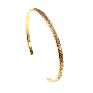 1 of 9: Close angled side view of a slim Nu Gold brass cuff bracelet standing upright on a white background. The image highlights the hand applied texture across the golden outer surface, the polished inner band, the curved profile, and one smooth open end.