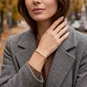 4 of 9: A woman in a gray herringbone coat and brown sweater wears a narrow golden brass cuff bracelet outdoors. The hand applied texture catches the light as her hand rests near her collar, while autumn trees and a city street form a softly blurred background.