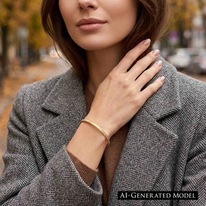 4 of 9: A woman in a gray herringbone coat and brown sweater wears a narrow golden brass cuff bracelet outdoors. The hand applied texture catches the light as her hand rests near her collar, while autumn trees and a city street form a softly blurred background. AI Generated Model.