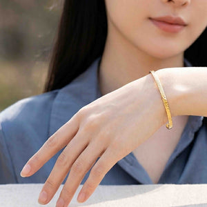 6 of 9: A woman in a slate blue blazer models a slim golden Nu Gold brass cuff bracelet on her extended wrist. The close view shows the bracelet’s narrow 3.5mm shape, polished edges, hand applied texture, and open cuff ends against a softly blurred indoor background.