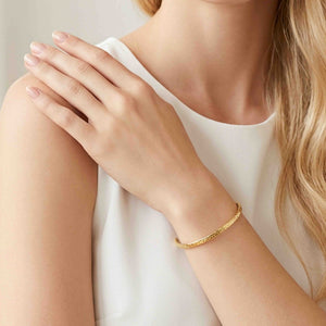 2 of 9: A blonde woman in a sleeveless white top wears a slim texturized Nu Gold brass cuff bracelet. Her hand rests gently on her opposite shoulder while the polished golden cuff circles her wrist, showing its narrow profile, textured outer surface, and open ends in soft neutral lighting.