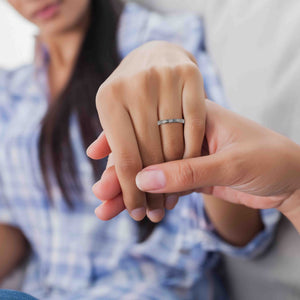 6 of 10: A close-up image of a woman wearing a slim bark-textured stainless steel ring on her ring finger. Another hand gently holds hers, suggesting a romantic or intimate moment. The woman is casually dressed in a plaid shirt, and the setting appears relaxed and cozy, possibly at home. The ring's polished finish and natural texture are clearly visible.