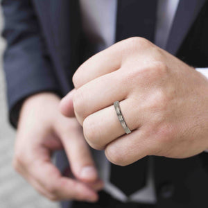2 of 10: A close-up image of a man wearing a slim stainless steel ring with a bark-textured finish on their ring finger. The man is dressed in a dark suit, suggesting a formal or professional setting. The ring has a clean, polished look and fits snugly on the finger.