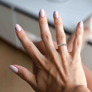 8 of 10: A close-up image of a woman's hand with manicured nails, showcasing a slim stainless steel ring with a bark-textured finish worn on the ring finger. The nails are painted in soft pink and silver tones, and the background suggests an indoor setting with natural light. The ring’s polished surface highlights its handcrafted, organic design.