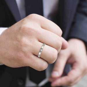 7 of 8: Close-up of a man wearing a 3mm flat stainless steel ring with a hand-chased texture on his ring finger, dressed in a dark suit and tie, suggesting a formal or professional setting.