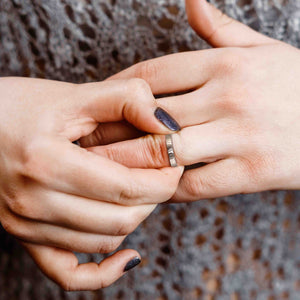4 of 8: Close-up of a woman's hand with glittery dark nail polish adjusting a 3mm flat stainless steel ring with a vertical hand-chased texture on their finger, with a lace-patterned top in the background.