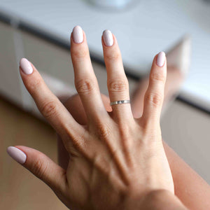 6 of 8: Close-up of a woman's hand wearing a 3mm flat stainless steel ring with a hand-chased texture on her ring finger, showcasing polished nails and a soft, neutral background.