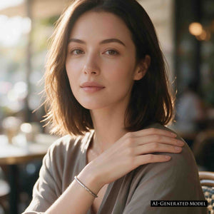 4 of 10: AI-generated female model wearing a 3mm Hammered Stainless Steel Cuff Bracelet by John S. Brana with a soft gray top in a warmly lit cafe. The slim, handcrafted bracelet is made from 316L surgical stainless steel and features a polished silver tone with an irregular hand hammered surface. Worn alone on her wrist, the narrow cuff creates a delicate metallic accent that complements relaxed contemporary women's fashion.