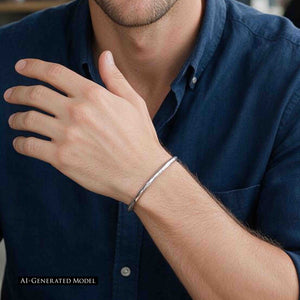 2 of 10: AI-generated male model wearing a 3mm Hammered Stainless Steel Cuff Bracelet by John S. Brana with a navy blue button-down shirt. The slender bracelet is handcrafted from 316L surgical stainless steel and features a reflective silver finish with subtle hammered impressions along the metal. Photographed close to the wrist, the minimalist open cuff demonstrates how narrow stainless steel jewelry complements casual men's clothing.
