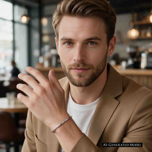 6 of 10: AI-generated male model wearing a 3mm Hammered Stainless Steel Cuff Bracelet by John S. Brana with a tan blazer and white shirt in a modern cafe. The slim, handcrafted 316L surgical stainless steel bracelet features a polished silver tone and hand hammered texture that catches the light. Styled as a minimalist men's fashion accessory, the narrow cuff adds a refined metallic accent to a smart casual outfit.
