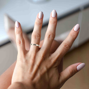 4 of 8: A Woman's Hand with a 3mm Hammered Stainless Steel Flat Ring and light pink nail polish on a blurred background