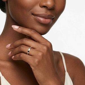 6 of 9: Close up of a woman wearing a slim polished stainless steel flat band ring on her finger, with her hand resting lightly under her chin. The bright silver ring has a narrow width, flat profile, and smooth reflective finish that stands out against her soft pink nails and neutral camisole. Its simple unisex style suits everyday wear, stacking, engagements, weddings, or anniversary gifting.
