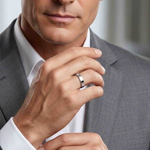 2 of 9: Close up of a man in a light gray suit and white dress shirt wearing a polished stainless steel flat band ring on his finger. His hand is raised near his chin, showing the ring’s slim width, flat profile, and bright mirror finish. The smooth silver tone adds a clean modern touch, making it well suited for everyday wear, weddings, engagements, or anniversary styling.