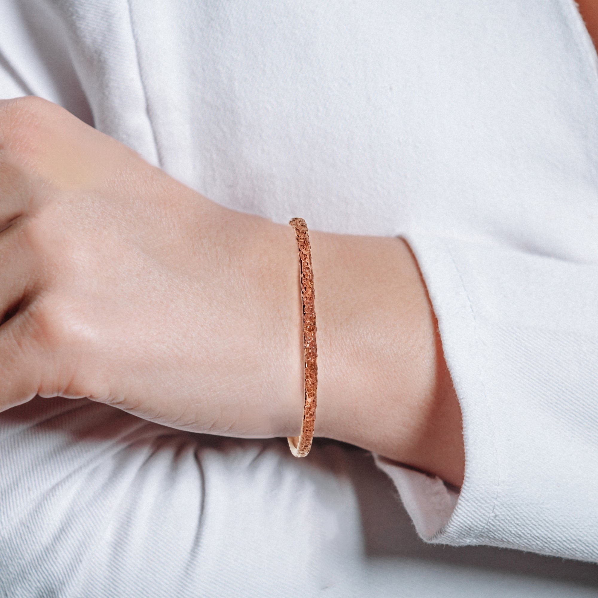 Close-up of a wrist wearing a slim textured copper cuff (≈3 mm wide). The narrow band shows a fine, granular hammered surface that catches light with a warm copper glow. Bracelet sits comfortably against the skin; a white long-sleeve top frames the wrist against a soft, neutral background.