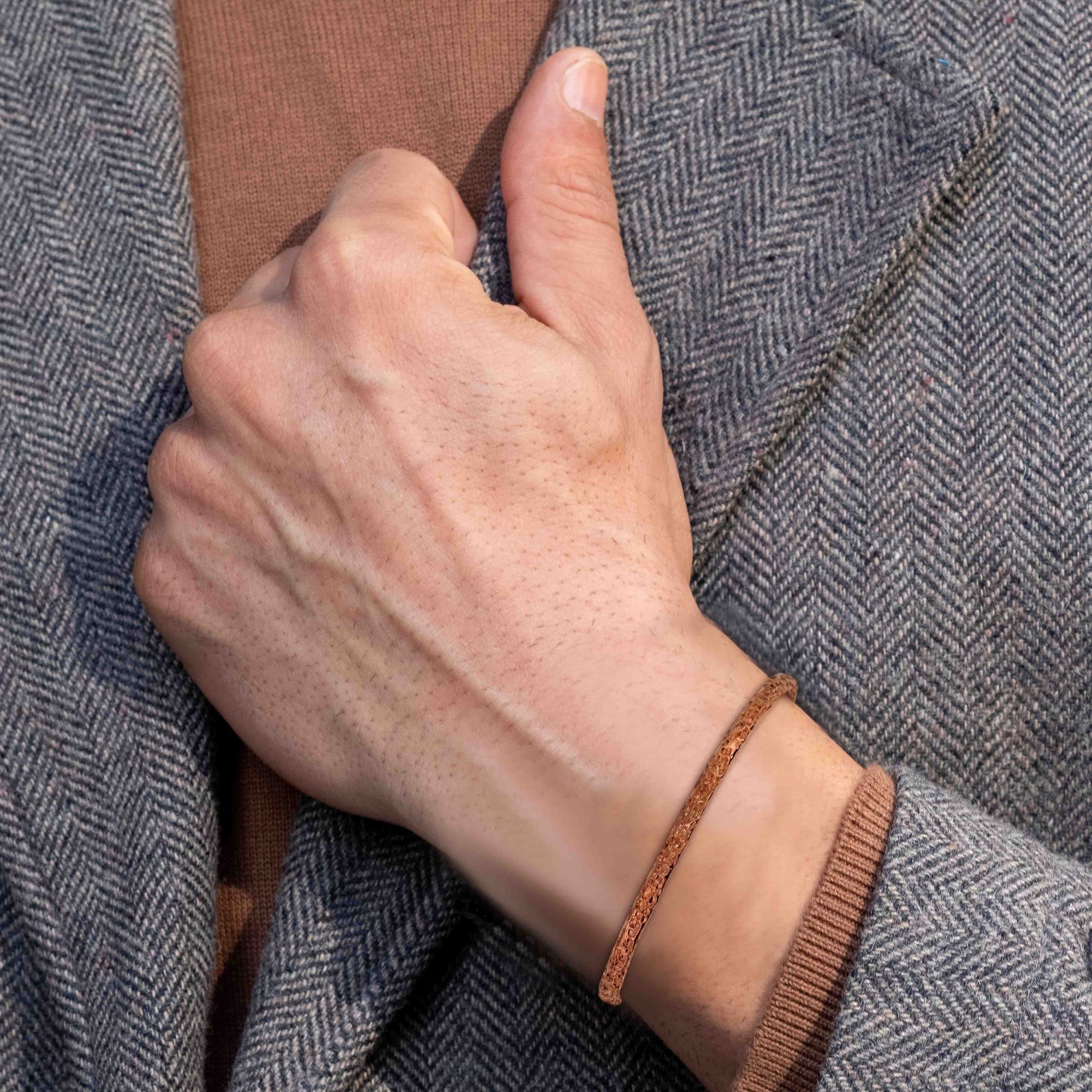 Close-up of a wrist wearing a very slim texturized copper cuff bracelet, styled with a gray herringbone jacket and warm brown knit underneath. The narrow solid copper cuff features a finely etched surface with subtle linear texture, giving it a matte, softly aged appearance rather than a high shine. The bracelet sits close to the wrist and blends naturally with the layered outfit, showing how the delicate copper band works as a minimal, understated accent for everyday wear.