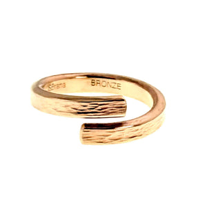 5 of 9: Angled top view of a handcrafted bronze bypass ring on a white background. The open band curves in a smooth wrap shape with two flat ends that nearly overlap. A hand applied bark texture runs across the outer surface, adding organic detail to the warm polished bronze finish. The inside of the band is stamped “BRONZE” and includes the maker’s mark.