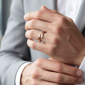 2 of 9: Close up of a hand wearing a slim bronze bypass ring on the ring finger. The open band wraps around with two straight ends that nearly meet, showing a subtle bark texture and warm coppery bronze shine. The ring is styled on a person in a light gray suit and white shirt, giving it a clean, modern, unisex look.
