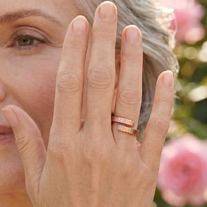8 of 9: Close view of a woman wearing a polished chased bronze bypass ring near her face in a garden setting. The open wrap ring has two textured ends with a small gap between them. Pink flowers and greenery appear softly blurred in the background.