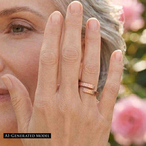6 of 9: Close view of a woman wearing a polished chased bronze bypass ring near her face in a garden setting. The open wrap ring has two textured ends with a small gap between them. Pink flowers and greenery appear softly blurred in the background.  AI Generated Model.