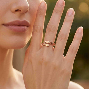 2 of 9: Close view of a woman wearing a slim chased bronze bypass ring on her finger outdoors. The two textured ends wrap around the finger with a small open space between them. Warm sunlight and a softly blurred garden appear in the background.