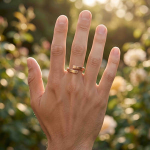 6 of 9: Close view of a hand wearing a polished chased bronze bypass ring outdoors. The ring has two narrow textured ends that overlap around the finger. Soft golden sunlight and blurred flowers create a warm garden setting.