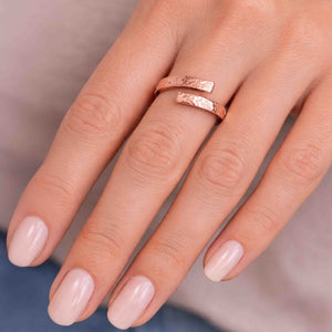 4 of 7: A close-up of a woman’s hand wearing a hammered copper bypass ring on the ring finger. The open-ended ring features two flat, textured copper tips that curve past each other, creating a modern wrap style. The warm rose-copper tone contrasts softly with smooth, light skin and pale pink manicured nails. The hammered finish reflects light, highlighting the handcrafted look against a neutral background.