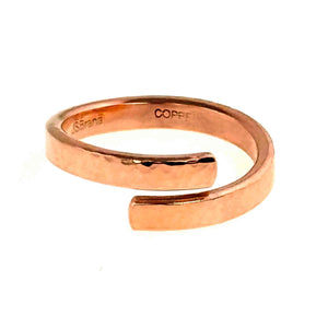 3 of 7: A handcrafted hammered copper bypass ring displayed against a white background. The ring features an open-ended design with smooth, squared tips that curve past each other in a wrap-around style. The surface has a lightly hammered texture and a polished finish, giving it a warm, reflective glow. Inside the band, the markings "J S Brana" and "COPPER" are visible, indicating the designer and material.