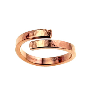 5 of 7: A handcrafted hammered copper bypass ring displayed on a white background. The ring has a warm reddish tone with a polished, reflective finish and a textured, hand-hammered surface. Its open-ended design features two squared tips that curve past each other, forming a wrap-around style. Inside the band, the inscriptions "J S Brana" and "COPPER" are clearly visible, indicating the artisan and metal type.