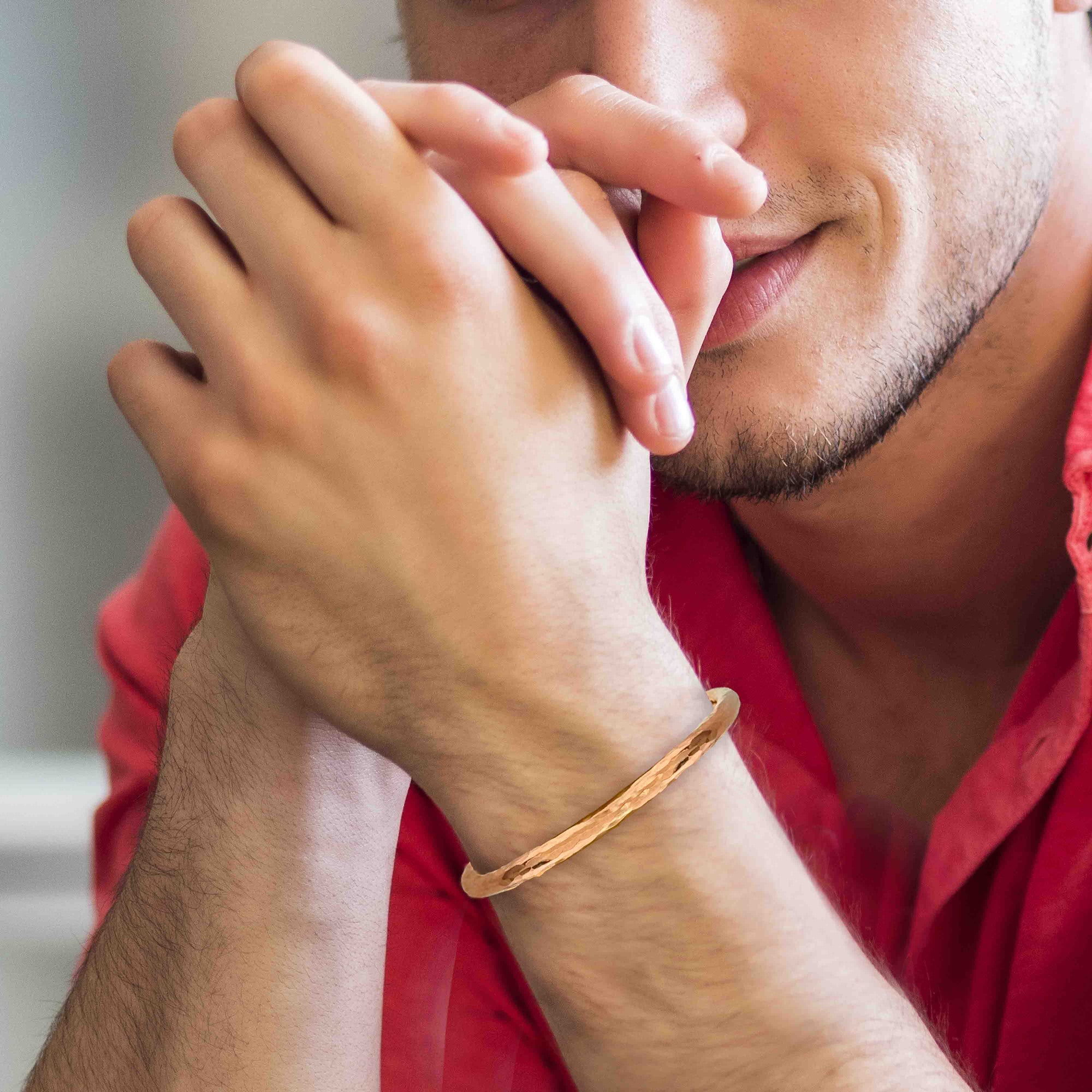 Close-up of a young man resting his clasped hands near his face, wearing a slim hammered copper cuff bracelet on his wrist. The bracelet has a warm, polished rose-gold tone with subtle faceted texture that catches the light, standing out against his light skin and casual red shirt. Soft, blurred background keeps the focus on the bracelet and shows how the narrow copper cuff looks in a relaxed, everyday styling on a man.