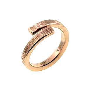 5 of 8: Angled view of the 4mm Linen Bronze Bypass Ring, highlighting its open wrap-around design and hand-applied linen texture. Made from raw, uncoated bronze with a warm, reddish tone, the ring features a lightly hammered grid-like pattern that adds rustic character. The inner band is engraved with “BRONZE” and “JSBrana,” indicating its handcrafted origin. A unisex piece perfect for everyday wear or as a meaningful anniversary gift.
