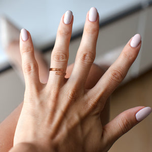 4 of 9: Woman’s hand wearing a copper bypass ring with a linen textured finish, shown with light neutral nail polish