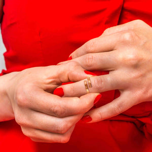 6 of 9: Hands with red nail polish adjusting a copper bypass ring with a linen textured finish against a red outfit background