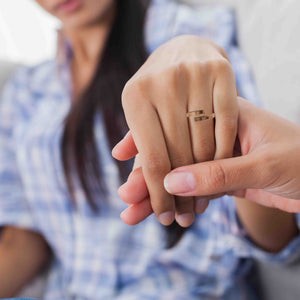 2 of 9: A close-up of a person gently holding a woman's hand, showcasing a 4mm matte bronze bypass ring on her finger. The ring features a brushed finish and open-ended design. The woman is wearing a blue plaid shirt, and the background is softly blurred.