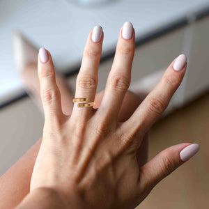 8 of 9: A close-up of a woman's hand wearing a 4mm matte bronze bypass ring on her middle finger. The ring features a brushed finish and open-ended design with squared edges. Her nails are manicured in soft pink shades, and the background is softly blurred, suggesting an indoor setting.