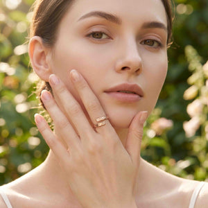 2 of 9: Close view of a woman wearing a warm bronze bypass ring on her finger while resting her hand against her face. Soft natural light and blurred green foliage highlight the ring’s open shape, polished finish, and textured ends.