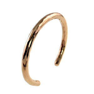 6 of 10: Angled left side view of the handmade 5.25mm hammered bronze cuff bracelet by John S. Brana against a clean white background. The polished solid bronze bracelet features a gently curved profile, reflective hand hammered surface, and smooth rounded ends. The photograph highlights the bracelet's thickness, warm golden coloring, and open cuff construction, showcasing its sculptural shape and craftsmanship.