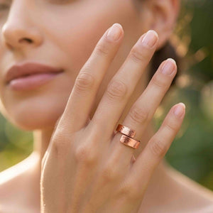 4 of 9: Hand wearing the 5.5mm bark copper bypass ring outdoors near soft greenery, showing the warm copper tone, textured bypass design, and polished shine on a natural feminine styling image.