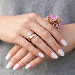 6 of 8: Close view of a woman wearing a chased copper bypass ring on her ring finger while holding small pink flowers. The warm copper band, open overlapping ends, and hand applied texture stand out against white nails and a soft gray background.