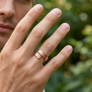 7 of 8: Close view of a man wearing a chased copper bypass ring outdoors. The open ring sits on the ring finger with overlapping textured ends, a polished warm copper finish, and a softly blurred green garden in the background.